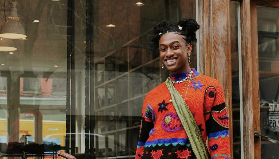 A smiling Black trans femme in their early 20s, wearing a red sweater, earrings, and necklaces.